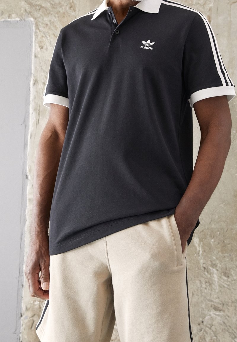 Svart Adidas-pikétröja med vit krage och ränder på ärmarna, bärs med beiga shorts, en hand i fickan mot en texturerad vägg.