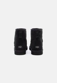 Botas de ante negras hasta el tobillo con puntera redondeada, suave forro textil, suela plana de goma y marca UGG en la parte posterior. Costuras decorativas a los lados.