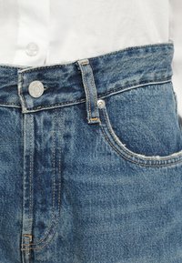 Blue denim jeans with a high waist, featuring a silver button closure, orange stitching, and a slight worn texture near the pockets.