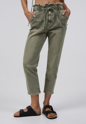 Persoon met olijfgroene paperbag cropped broek en zwarte slip-on sandalen met kurkzool, staand met handen in de zakken.