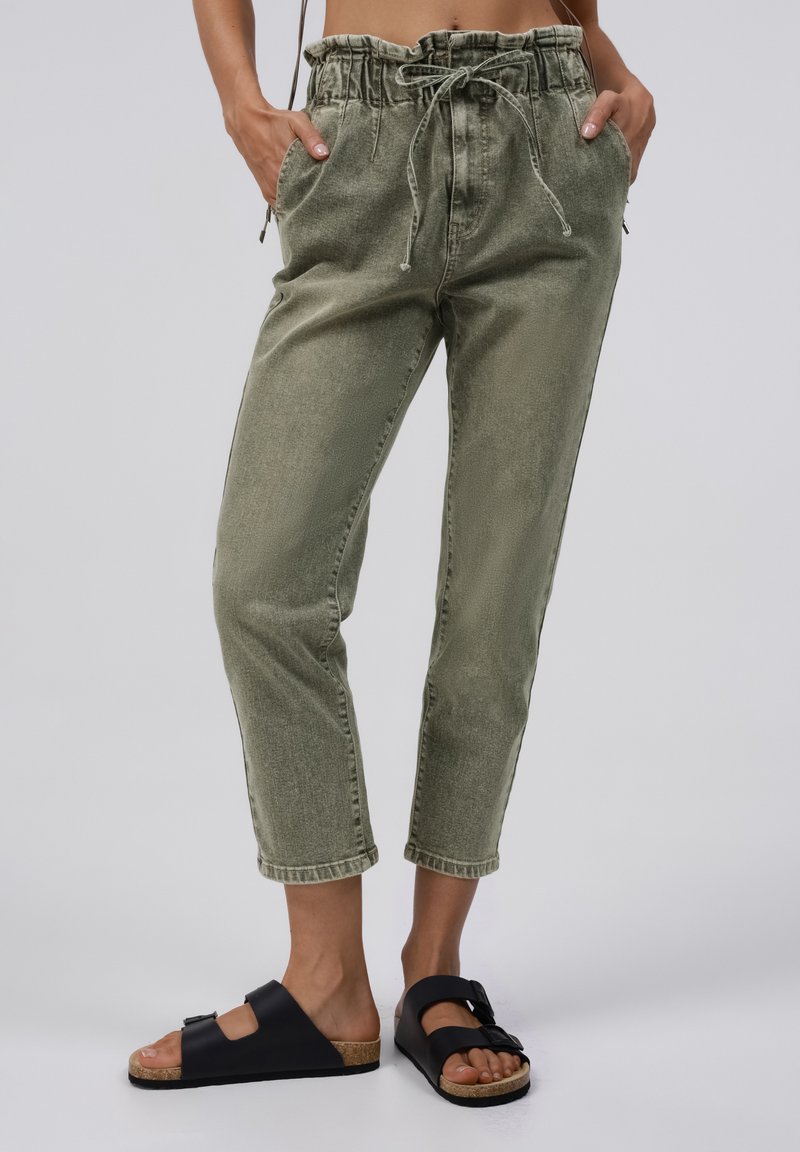 Personne portant un pantalon court taille paperbag vert olive et des sandales noires à enfiler avec semelles en liège, debout les mains dans les poches.