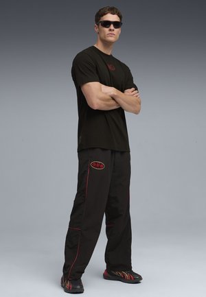 Mand iført sorte solbriller, sort skjorte med rød logo, sorte bukser med røde detaljer og sorte sneakers med røde og gule accenter, stående med armene over kors.