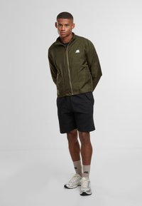 Mladý muž stojící s rukama za zády, oblečený v olivově zelené bundě, černých šortkách, béžových ponožkách a stříbrných běžeckých botách.