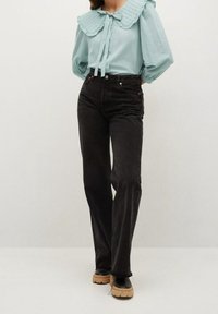 Donna che indossa una blusa azzurro chiaro con un ampio colletto arrotondato e un fiocco, jeans neri a vita alta dritti e scarpe nere con suole spesse beige.
