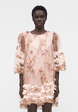 Γυναίκα με διάφανο ροζ φόρεμα με κεντήματα λουλουδιών και εφαρμογές 3D λουλουδιών, στέκεται μπροστά σε απλό λευκό φόντο.