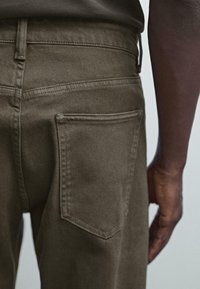 Close-up of a person wearing olive-green pants showing the back pocket and part of their right arm against a plain background.