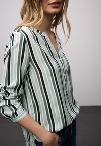 Femme portant une chemise à manches longues à rayures verticales vertes, blanches et noires, avec des boutons et une main dans la poche d'un jean bleu.