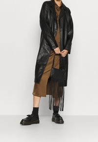 Czarny skórzany długi płaszcz z wykładanym kołnierzem, w połączeniu z brązową sukienką zapinaną na guziki. Zawiera czarną torbę z frędzlami. Ubrano czarne buty.