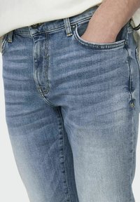 Lichtblauwe denimjeans met een licht vervaagde textuur, voorzien van een traditionele vijf-pocketontwerp en metalen accenten bij de taille en zakken.