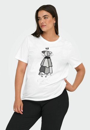 Femme aux cheveux foncés portant un t-shirt blanc avec un croquis de mode en noir et blanc, debout, une main sur la hanche, sur un fond uni.