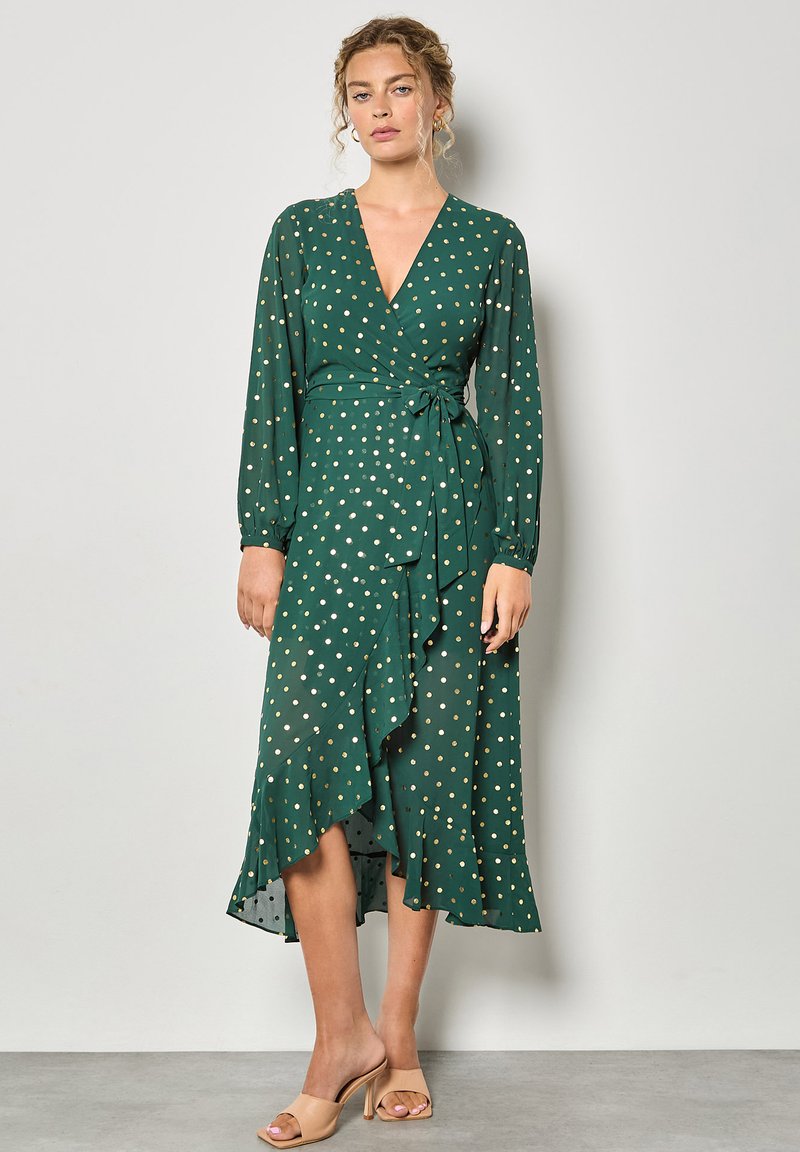 Abito avvolgente verde con pois bianchi e dorati, maniche lunghe, orlo arricciato e dettaglio in vita con fiocco, realizzato in tessuto leggero.