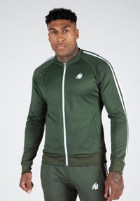 Grüne sportliche Zip-Jacke mit weißen Streifen an den Schultern, ausgestattet mit einem Frontreißverschluss, gerippten Bündchen und einem Logo auf der Brust.