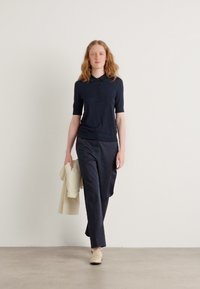 Cămașă polo cu mânecă scurtă de culoare bleumarin și pantaloni închiși la culoare, asortați cu o jachetă de nuanță deschisă așezată pe un braț. Design simplu și nuanțe neutre.