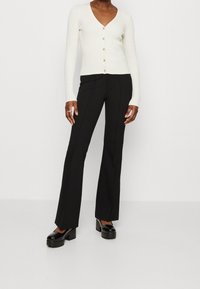 Cardigan blanco de manga larga con relieve y pequeños botones, combinado con pantalones negros de campana y zapatos de plataforma negros gruesos.