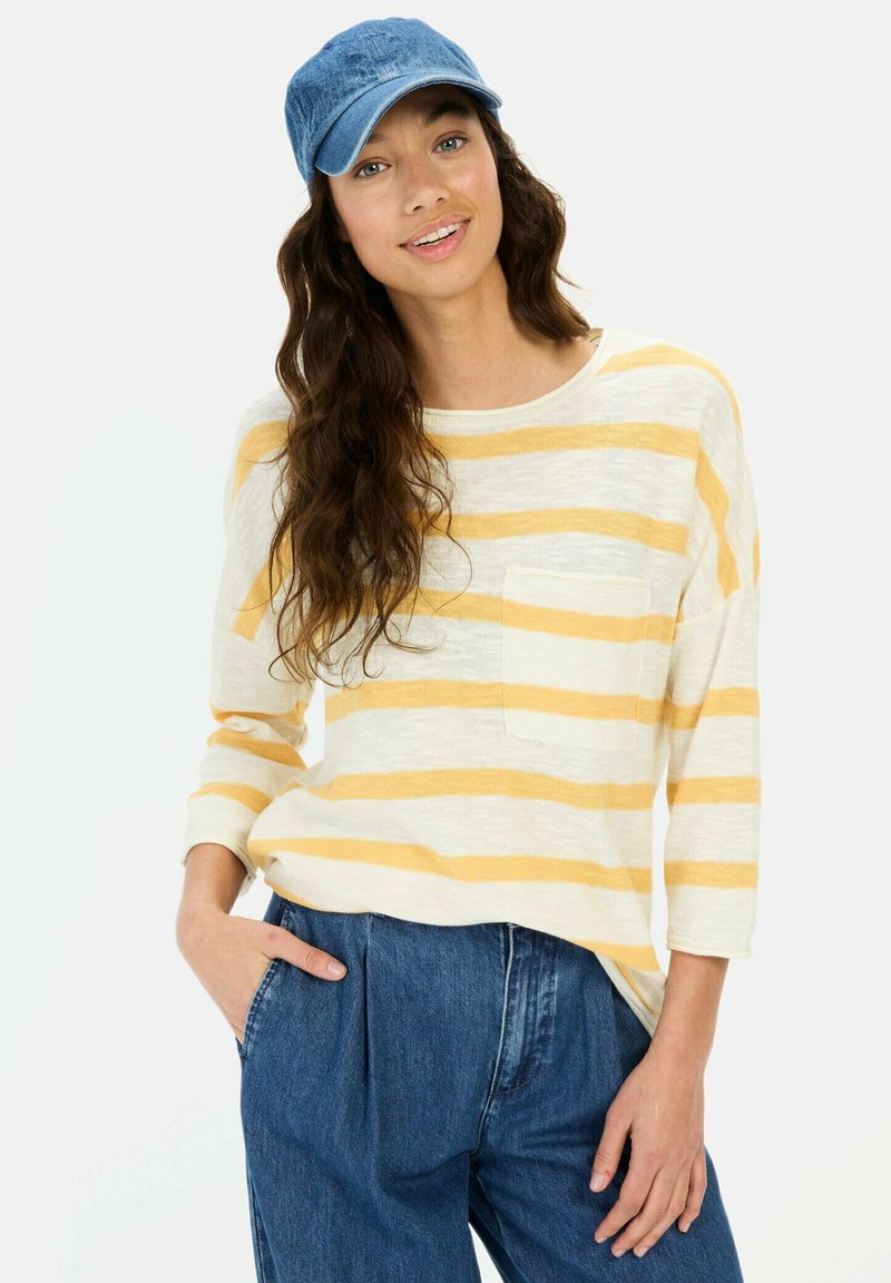 Gestreiftes Langarmhemd in Creme und Gelb mit einer Tasche; kombiniert mit blauen Jeans und einer hellblauen Denim-Mütze.