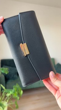 Portofel negru texturat, fabricat dintr-un material asemănător pielii, cu un închizător de culoare aurie și margini cusute cu atenție. Ținut într-o mână, împotriva unui fundal neclar.