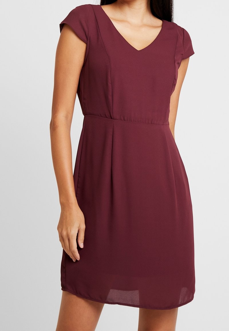Femme portant une robe bordeaux à manches courtes, longueur genou, col en V et taille cintrée sur un fond uni.