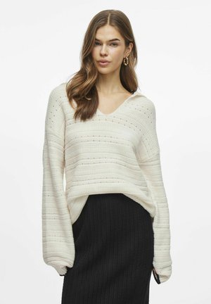 Biały sweter dzianinowy z dekoltem w serek, z poziomymi paskami i małymi akcentami. Długie rękawy i luźny krój, zestawiony z czarną spódnicą z prążkowanej dzianiny.