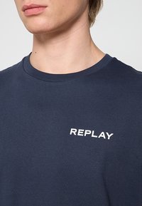 Jeune personne portant un t-shirt bleu marine à col rond avec le texte blanc "REPLAY" imprimé sur la poitrine.