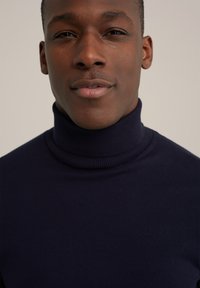 Sötét tengerészkék garbós pulóver sima textúrával és bordás nyakkal, rövid hajú személyen, aki semleges arckifejezéssel viseli.