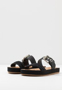 Black sandals with woven straps, cork insoles, and textured rubber soles. Features a silver buckle and rubber accents.
