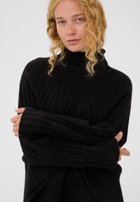 Šviesiaplaukė moteris su garbanotais plaukais, vilkinti juodą raukšlėtą golfą, su sudėtomis rankomis, prieš paprastą šviesią fono.