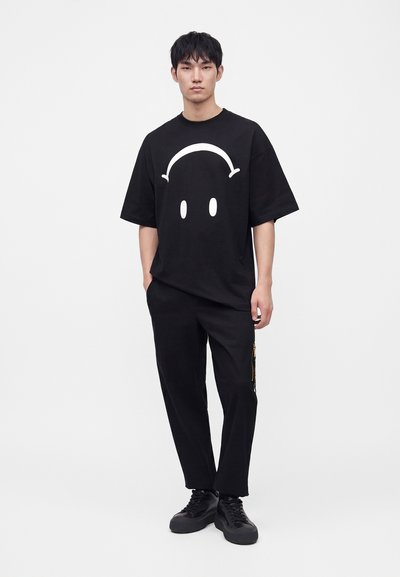 Jeune homme debout portant un t-shirt noir oversize avec un motif abstrait blanc représentant un visage, un pantalon noir et des baskets noires sur un fond uni.