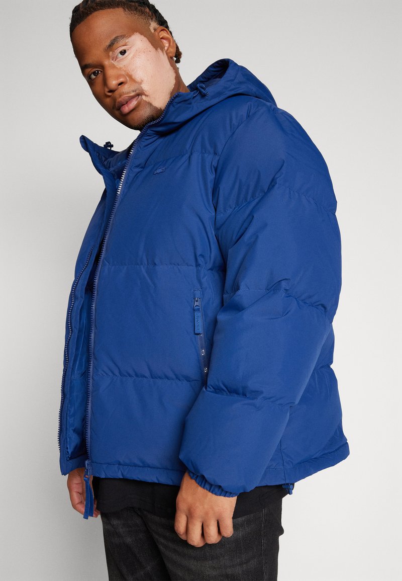 Giacca a vento blu con cappuccio, dotata di chiusura con zip e polsini elastici. Realizzata in materiale lucido, design trapuntato e un piccolo logo sul petto.