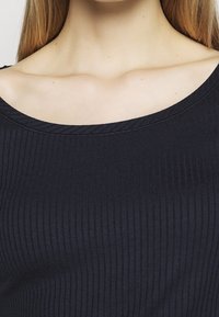 Nahaufnahme des Nackens und der Schlüsselbeine einer Frau, die ein schwarzes, geripptes Boot-Ausschnitt-Top trägt und gerade hellbraune Haare hat.