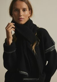 Schwarzer Strickpullover mit einem großen, gewickelten Schal. Das Design umfasst eine gerippte Textur und kontrastierende graue Nähte an den Ärmeln.