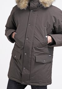 Giacca parka verde oliva scuro con cappuccio foderato in pelliccia. Presenta bottoni a pressione, due tasche frontali e una texture trapuntata.