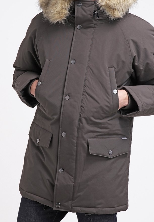 Giacca parka verde oliva scuro con cappuccio foderato in pelliccia. Presenta bottoni a pressione, due tasche frontali e una texture trapuntata.