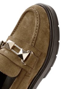 Mocassin en daim marron avec semelle noire, décoré d'une chaîne métallique argentée en forme de pyramide sur le dessus.