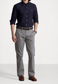 Marinblå skjorta med knappar och en liten röd logga, ljusgrå byxor, vävd brun bälte och bruna loafers med snören. Enkelt och klassiskt design.