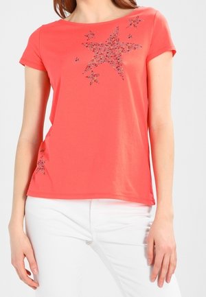 Kobieta ubrana w koralowy top z krótkim rękawem z motywem kwiatowych gwiazd, w połączeniu z białymi spodniami, stojąca na jednolitym tle.