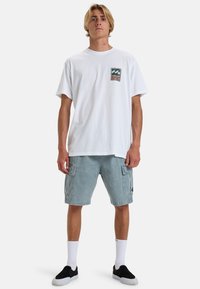 White cotton t-shirt with a small multicolored graphic on the left chest, paired with light blue cargo shorts and black slip-on shoes.