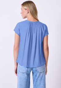 Blusa blu a maniche corte con una texture morbida e liscia. Presenta dettagli gathered sul retro e una vestibilità ampia. Abbinata a jeans blu chiaro.