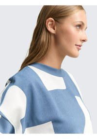 Blauwe sweatshirt met grote witte geometrische patronen, gemaakt van zachte stof met een ronde hals en een ontspannen pasvorm. Soepele textuur en casual ontwerp.