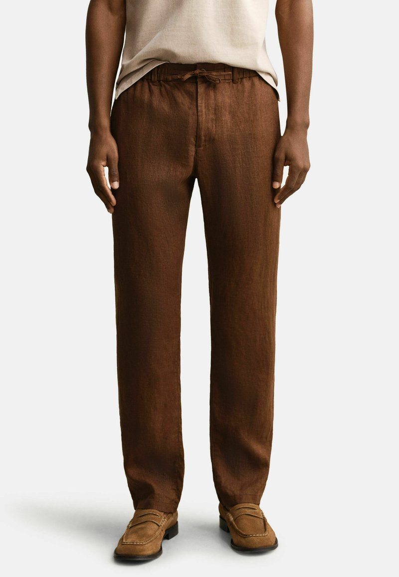 Mann trägt braune locker sitzende Leinenhose mit Bindeband in der Taille und hellbraune Wildleder-Loafer, stehend vor einem schlichten weißen Hintergrund.