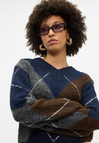 Sweter dzianinowy z dekoltem w serek, ozdobiony diamentowym wzorem w kolorach granatowym, brązowym i szarym, z białymi akcentami w przeszyciach oraz miękką fakturą.