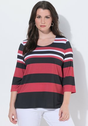 Donna che indossa una maglia a maniche lunghe con strisce orizzontali nere, rosse, bianche e rosa, abbinata a pantaloni bianchi, in piedi davanti a uno sfondo bianco.