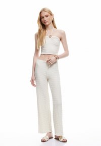 Kvinde med langt blont hår iført en tekstureret hvid strapless crop top, matchende vide bukser, guldringe, armbånd og cremede slidesandaler.