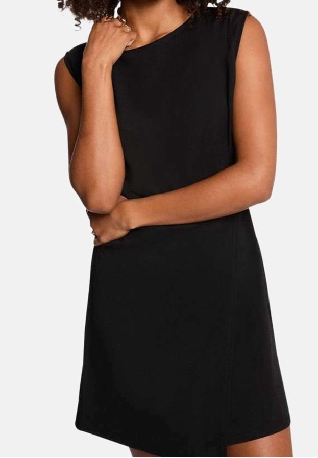 Femme aux cheveux bouclés portant une robe noire sans manches, un bras croisé sur la taille et l'autre main près du menton.