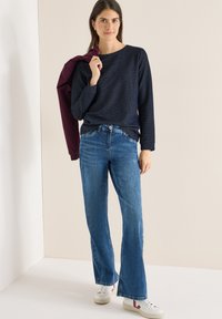 Granatowy top z długim rękawem o teksturowanym wzorze, zestawiony z jasnoniebieskimi dzwonami i białymi trampkami z bordowymi akcentami. Kurtka zarzucona na ramię.