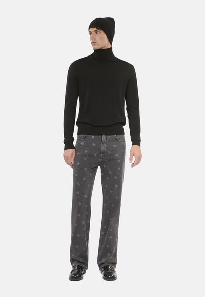 Maglione a collo alto nero, aderente. Jeans grigi con motivo a stelle. Scarpe nere. Modello in posa rivolto a sinistra su uno sfondo bianco.