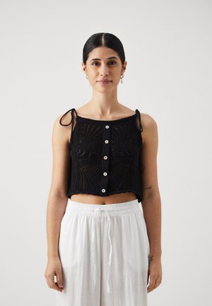 Black knitted crop top with openwork pattern, narrow shoulder ties, and white buttons down the front. Paired with white elastic-waist pants.