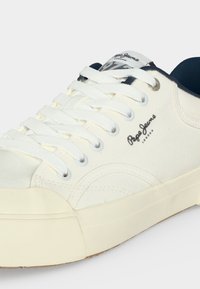 Baskets en toile blanche avec lacets blancs, semelle en caoutchouc crème, intérieur bleu marine et logo Pepe Jeans London sur le côté et la languette.