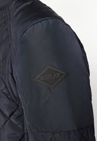 Geacă căptușită de culoare bleumarin, cu un model matlasat pe umeri. Prezintă o etichetă rectanguală cu logo-ul „REPLAY” de culoare neagră. Textură netedă, formă ajustată.