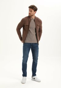 Cardigan marrone in maglia con zip, texture a coste, due tasche sul petto, maglietta grigia a girocollo, jeans di denim blu e sneakers bianche.