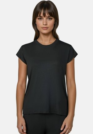 Vrouw met stijl bruin haar die een effen zwart T-shirt met korte mouwen draagt, staand tegen een lichtgrijze achtergrond.